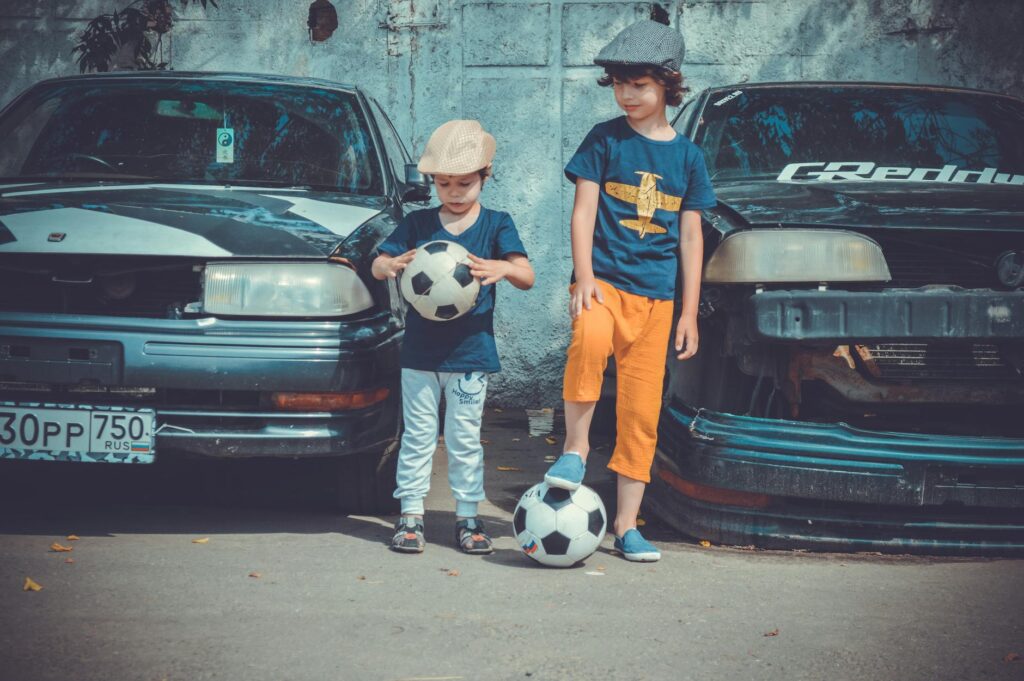 two boys playing soccer ball beside cars